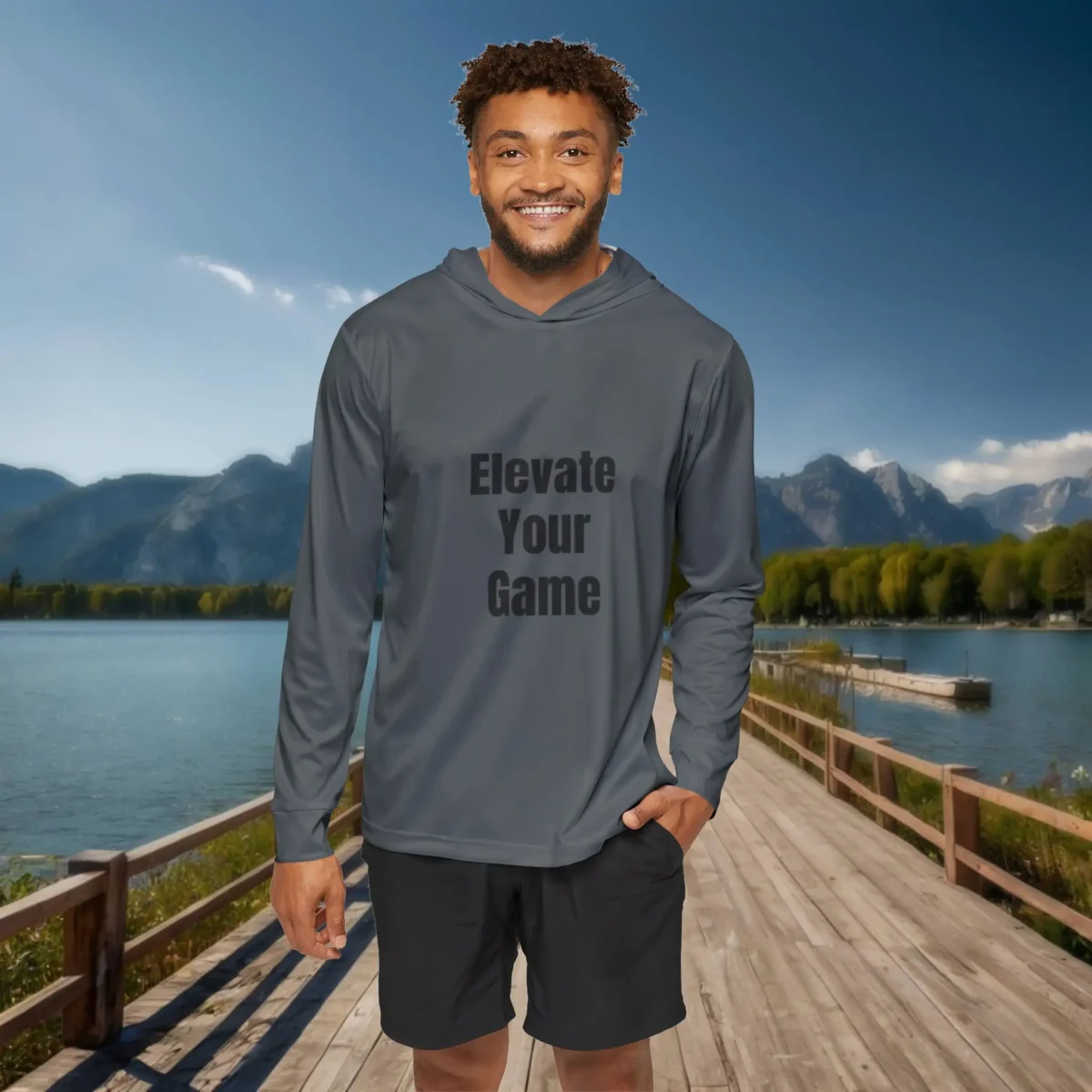 Smiling man in a gray hoodie with the motivational slogan “Elevate Your Game” standing on a wooden boardwalk by a scenic lake and mountains.