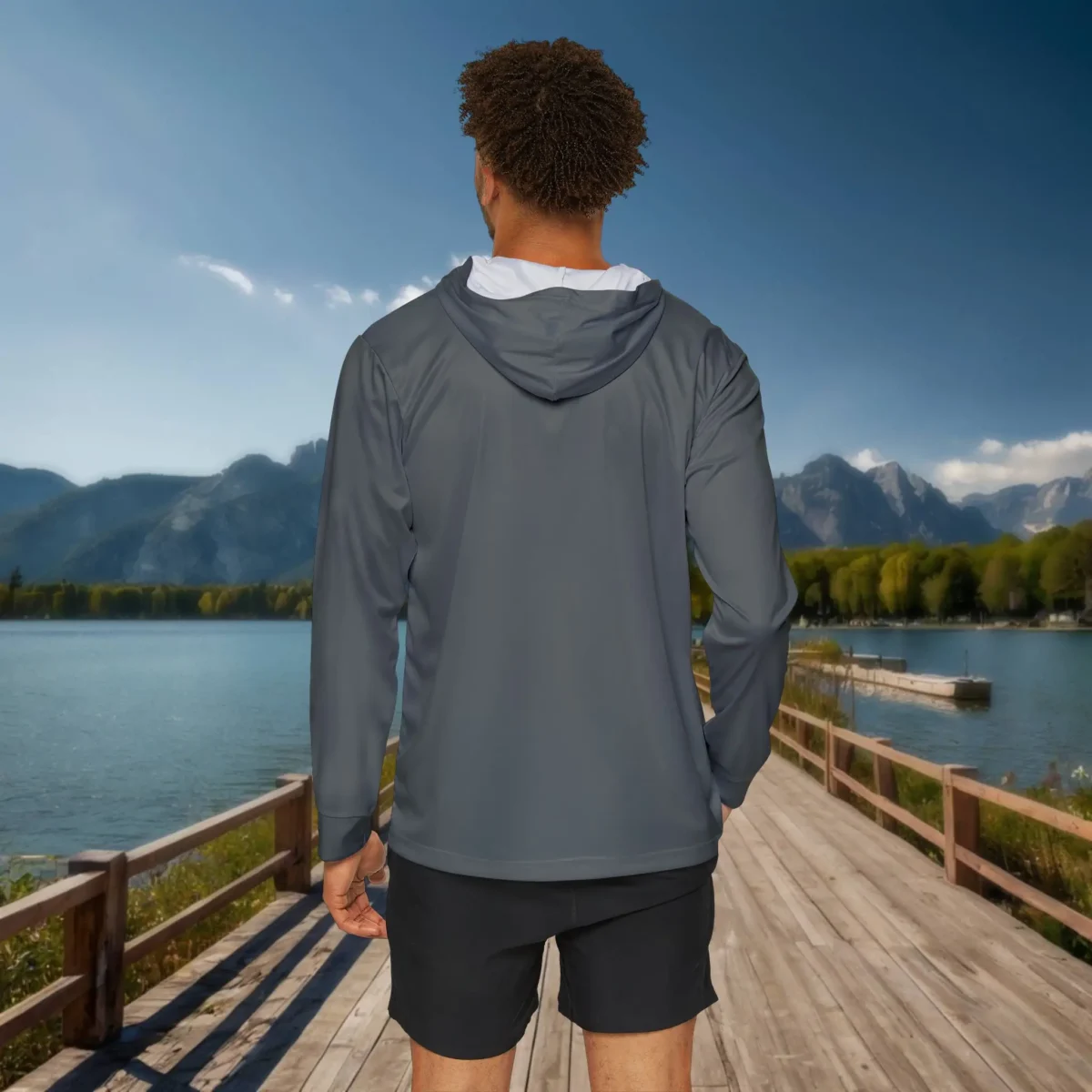 Back view of a man in a gray hoodie standing on a wooden dock with lake and mountain scenery.