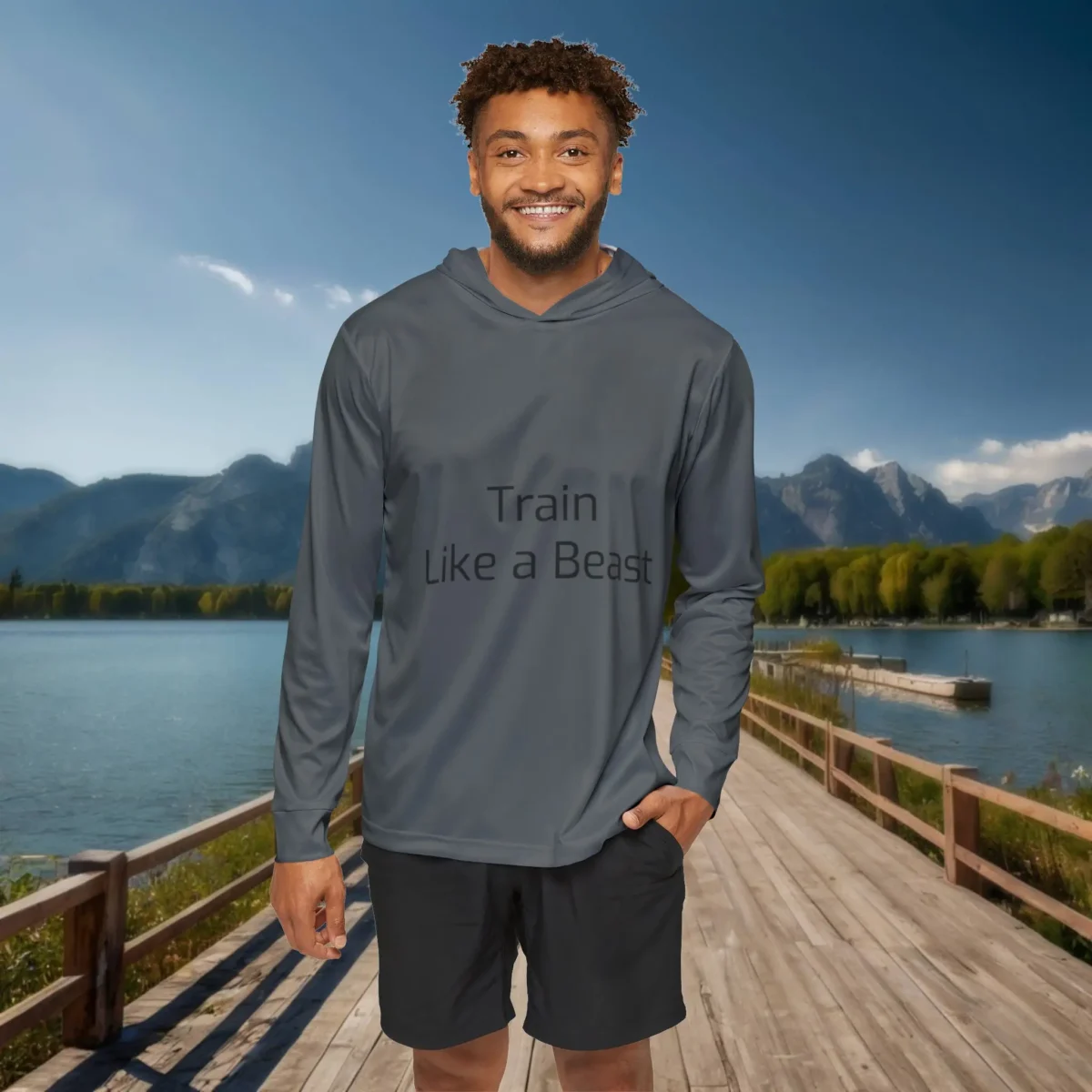 Man wearing a gray hoodie with the text “Train Like a Beast” on the front, standing on a lakeside boardwalk with mountains in the background.