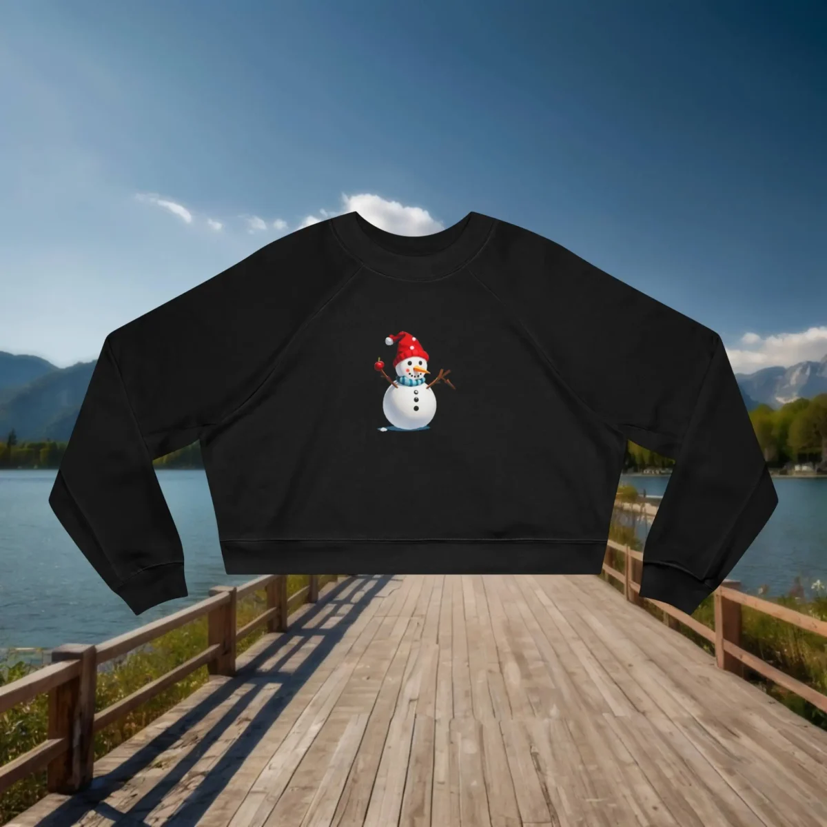 Black cropped sweatshirt with a cartoon snowman design laid flat on a dock by a scenic lake and mountain background.