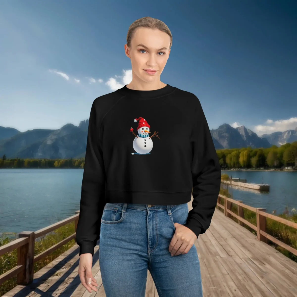 Woman wearing a black cropped sweatshirt with a cheerful snowman design, standing on a wooden dock with a lake and snowy mountain backdrop.