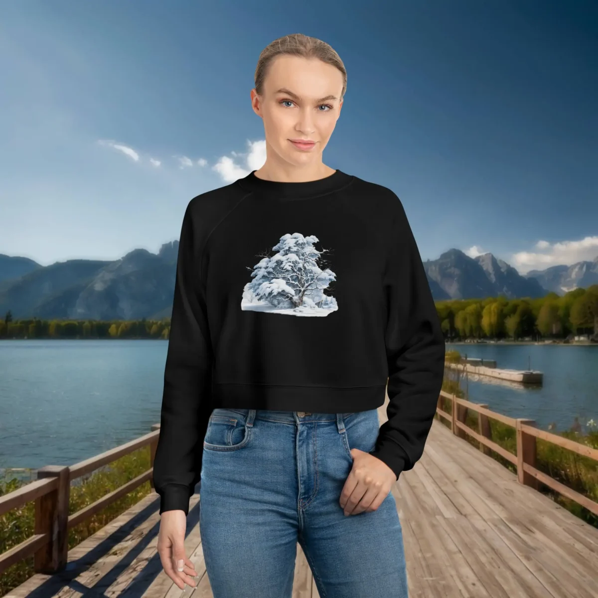 Woman wearing a black cropped sweatshirt with a snowy tree design, standing on a dock by a lake with mountains in the background.
