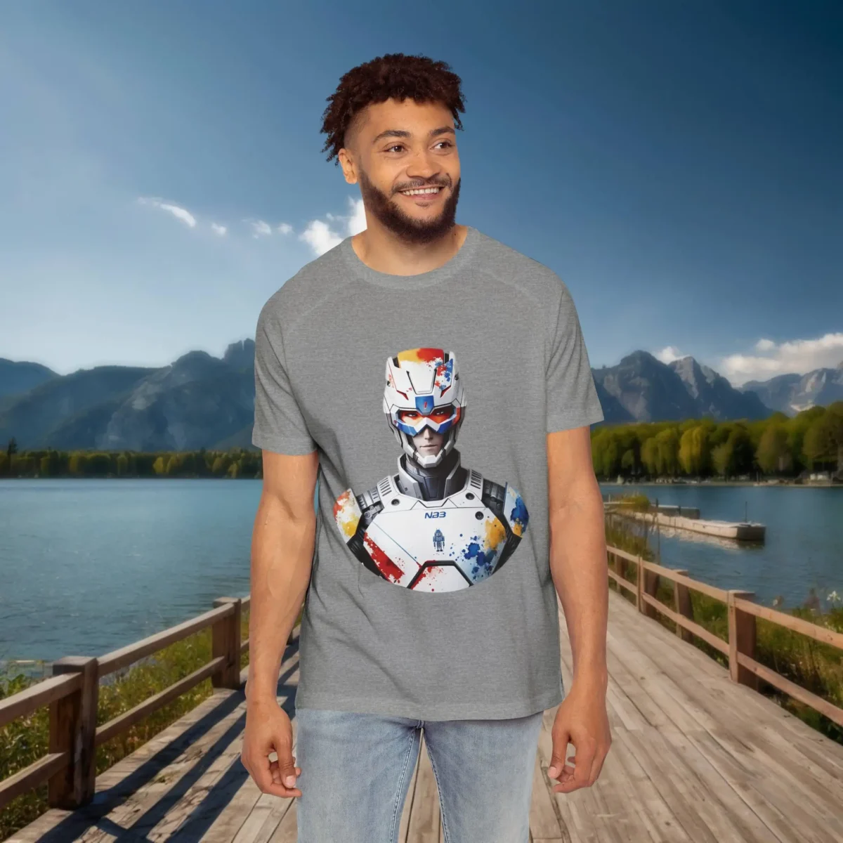 Man wearing a grey raglan t-shirt with a colorful futuristic soldier graphic, standing on a wooden dock by a lake with mountains in the background.