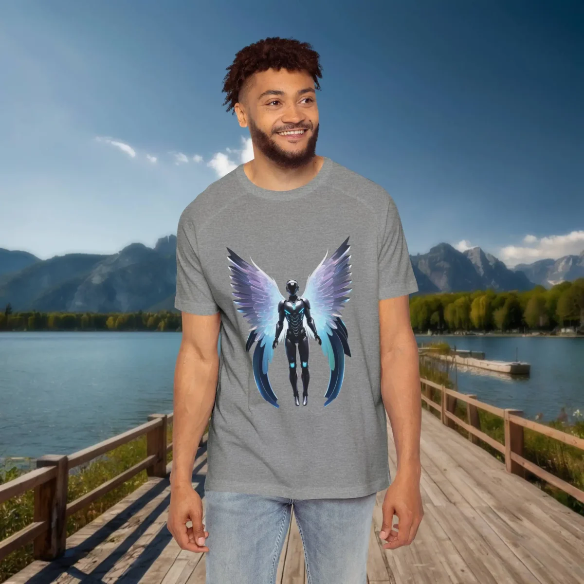 Man wearing a grey raglan t-shirt with a futuristic humanoid figure featuring glowing wings, standing on a dock by a lake with mountains in the background.