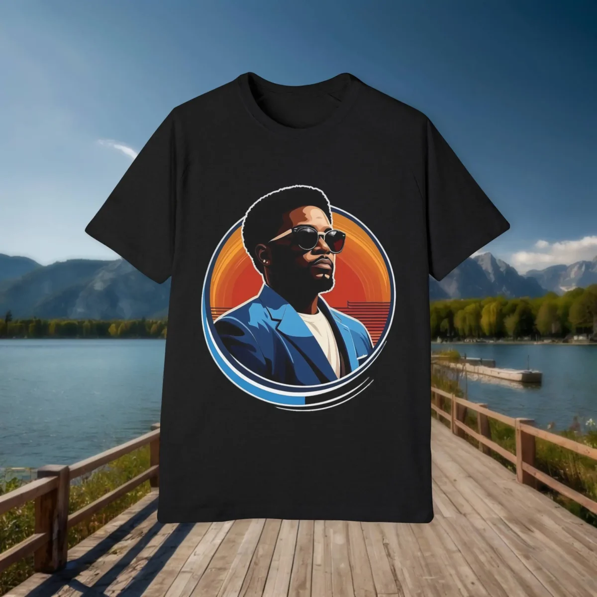 Black t-shirt laid flat with a stylized circular portrait of a man wearing sunglasses and a blue blazer, shown on a dock with a lake and mountains in the background.