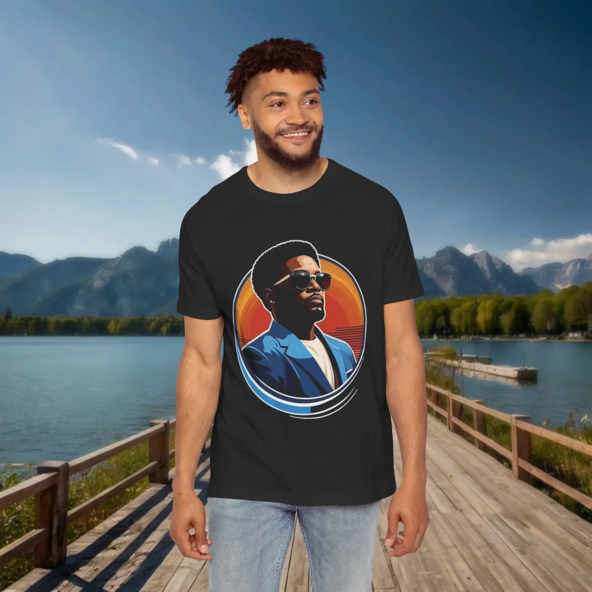 Man wearing a black raglan t-shirt with a colorful graphic of a stylish man in sunglasses and a blazer, standing by a scenic mountain lake.