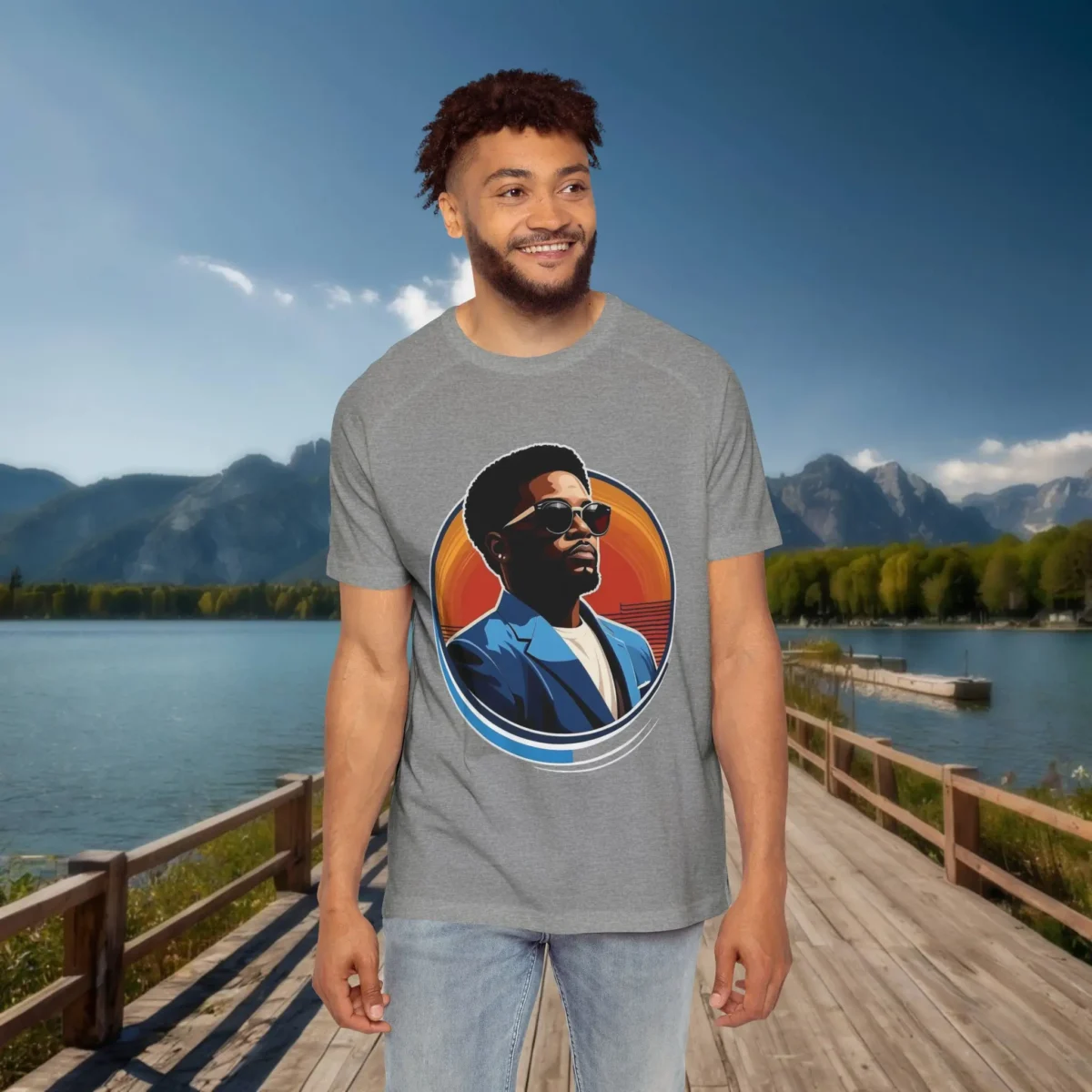 Man wearing a grey raglan t-shirt featuring a stylized portrait of a man in sunglasses and a blue blazer, standing on a dock with a lake and mountain background.