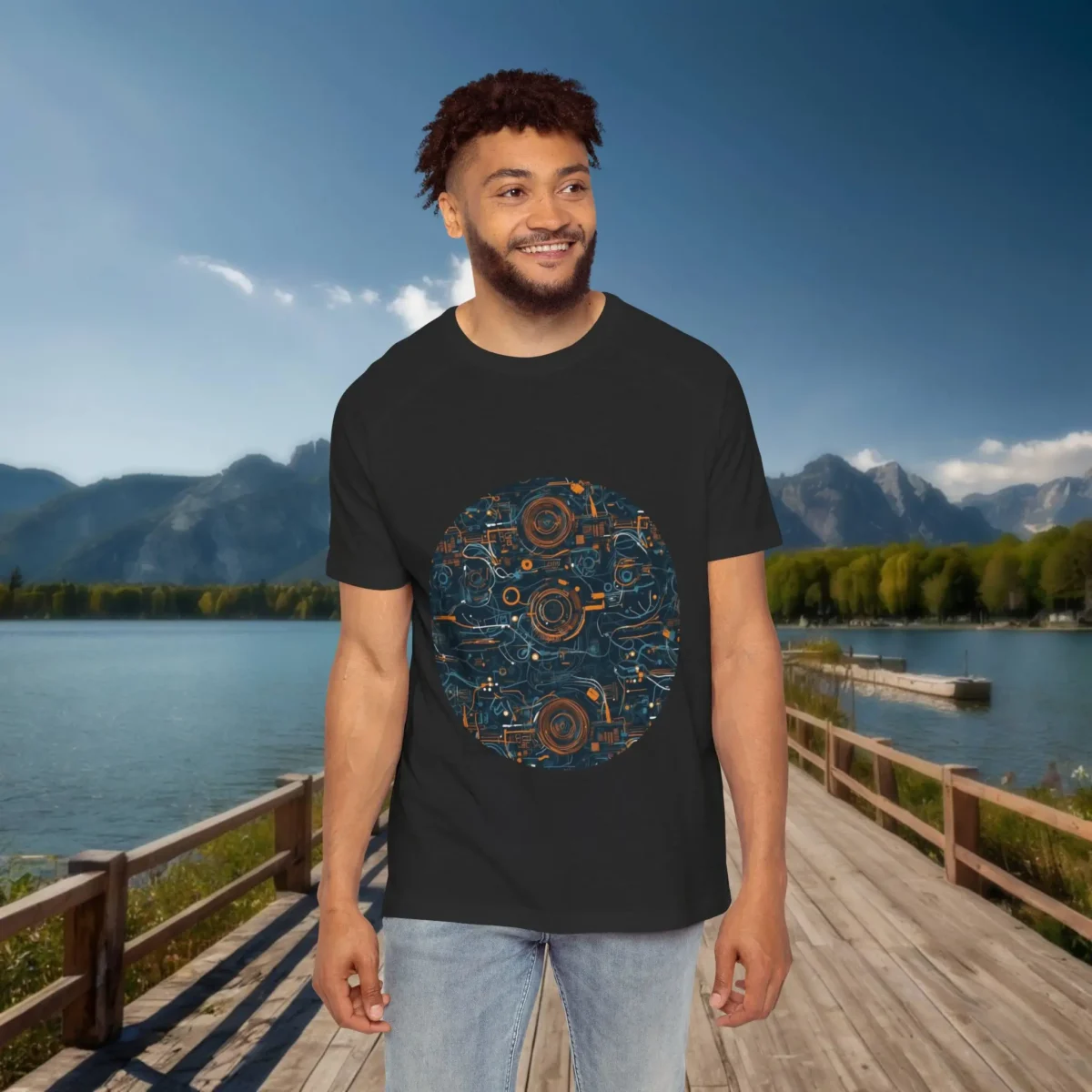 Man wearing a black raglan t-shirt with a circular mechanical gear and circuit pattern, standing on a wooden dock with a lake and mountains in the background.