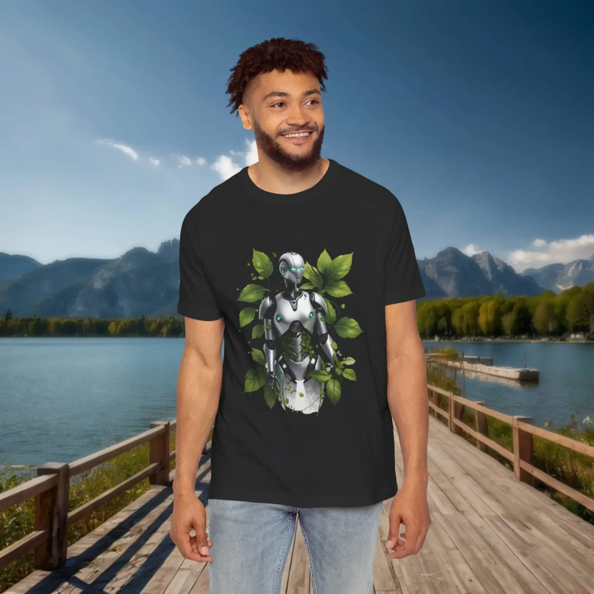 Man wearing a black raglan t-shirt with a nature-robot fusion design, showing a silver humanoid figure surrounded by leaves, standing by a lake with mountains.