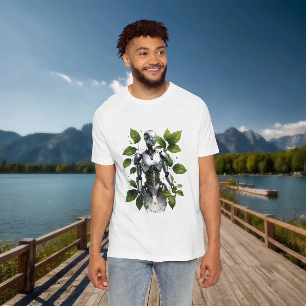Man wearing a white raglan t-shirt featuring a silver humanoid robot surrounded by green leaves, standing on a dock with a scenic lake and mountain backdrop.