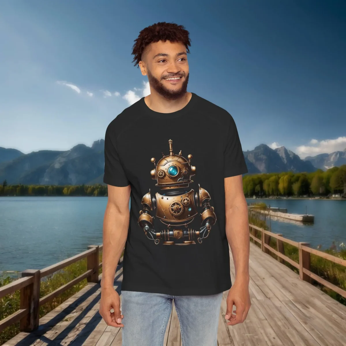 Man wearing a black raglan t-shirt featuring a bronze vintage robot with blue glowing eye and gears, standing outdoors on a lakeside wooden dock with mountains in the background.
