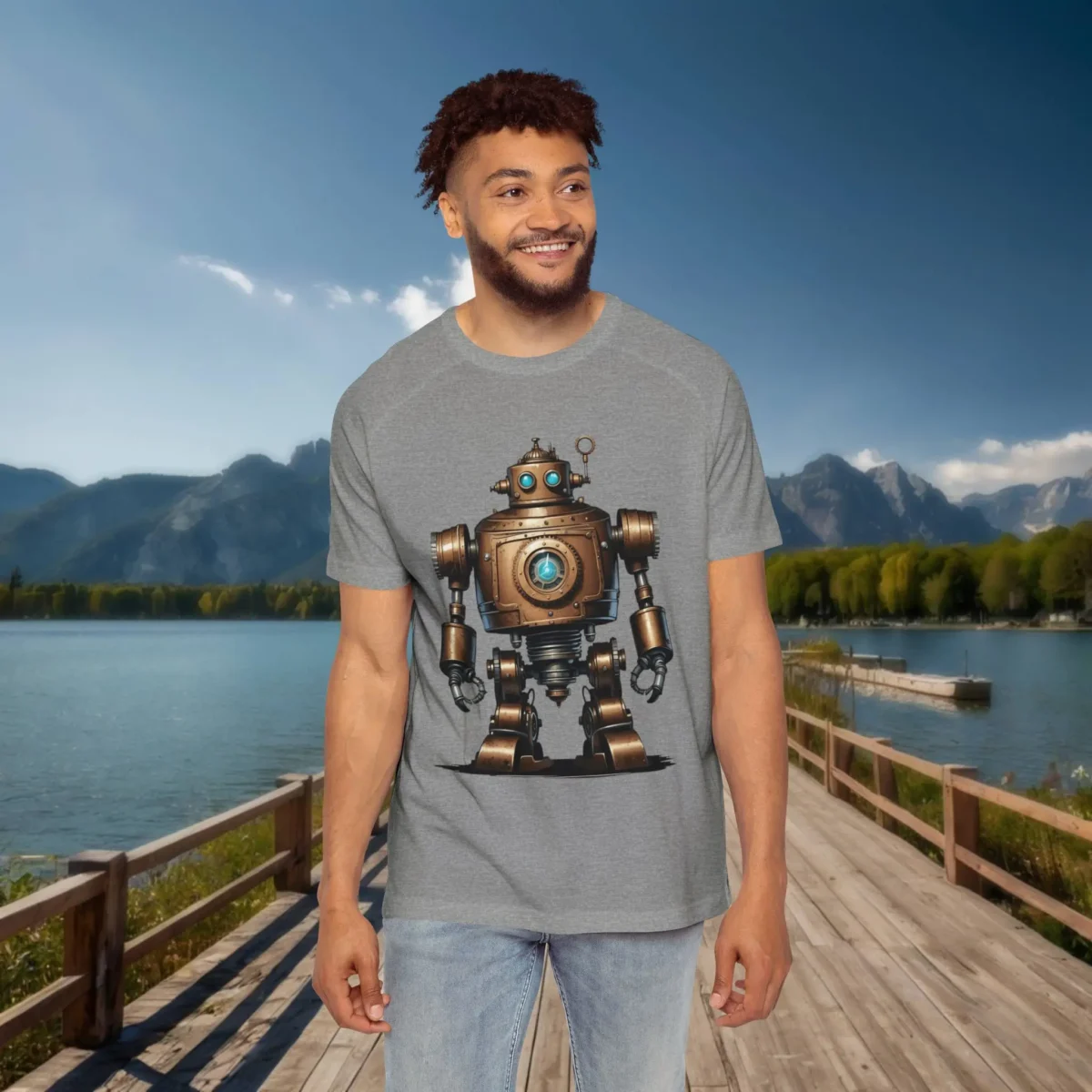 Man wearing a grey raglan t-shirt with a detailed vintage bronze robot design, standing on a wooden dock with a scenic background of lake and mountains.