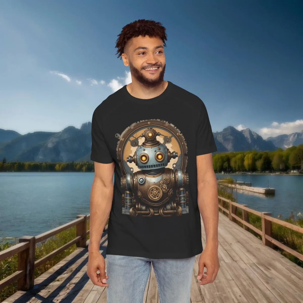 Man wearing a black raglan t-shirt featuring a cute vintage-style steampunk robot with glowing orange eyes and metallic gears, standing on a dock with lake and mountain scenery.