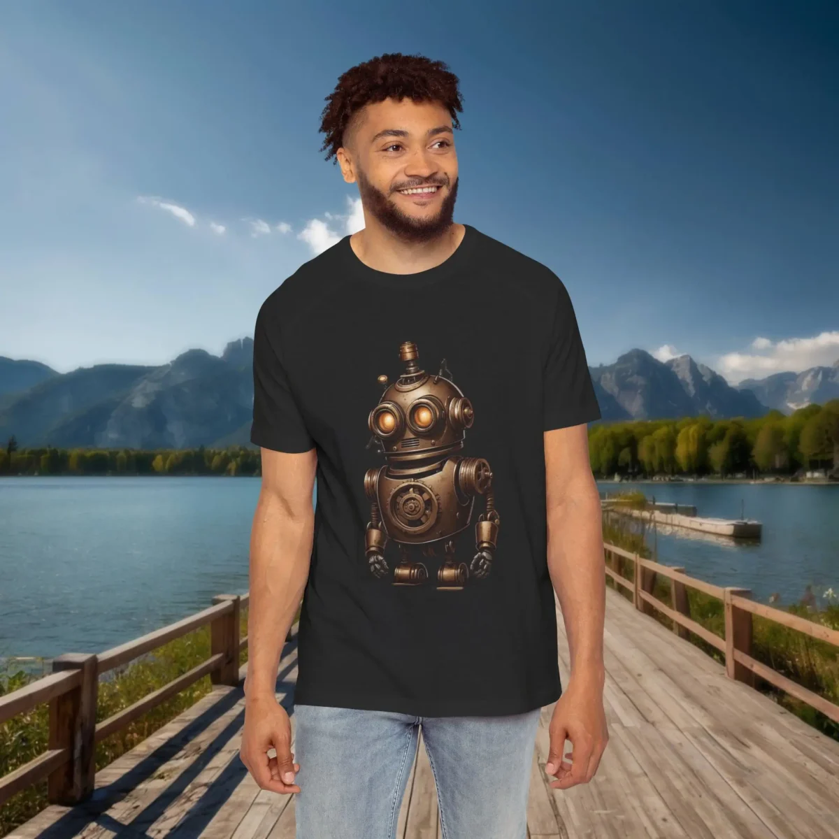 Man wearing a black raglan t-shirt with a bronze vintage-style robot illustration featuring gears and glowing orange eyes, posed on a wooden dock by a lake with mountain scenery.
