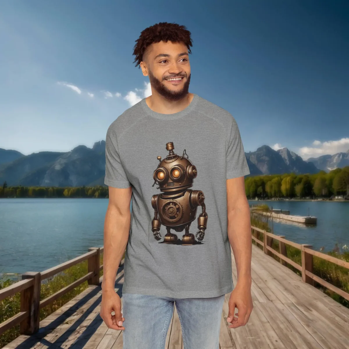 Man wearing a grey raglan t-shirt featuring a cute bronze vintage robot with glowing orange eyes and industrial details, standing on a dock in front of a lake and mountains.