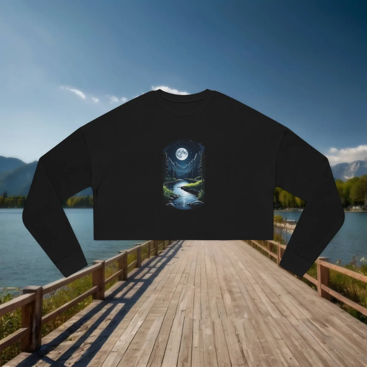 Black cropped sweatshirt with a moonlit river and forest design laid flat on a wooden dock over a serene mountain lake.