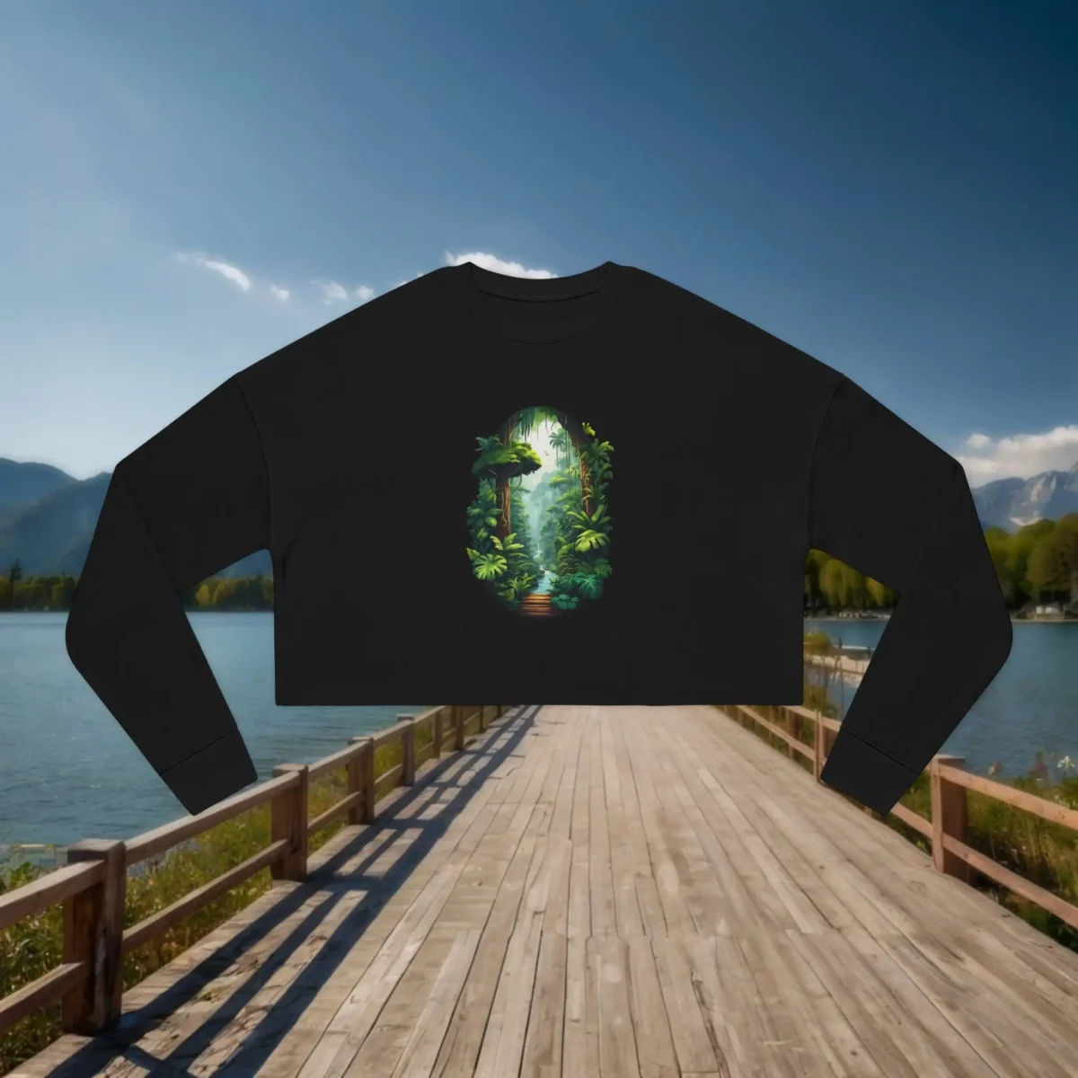 Black cropped sweatshirt with a detailed rainforest path and canopy print laid flat on a wooden dock over a mountain lake.