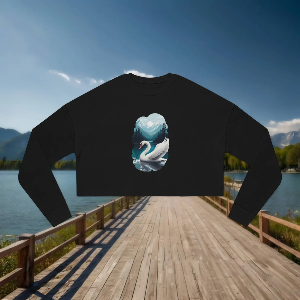 Black cropped sweatshirt with a swan and mountain lake design laid flat on a wooden dock overlooking a calm lake and mountains.