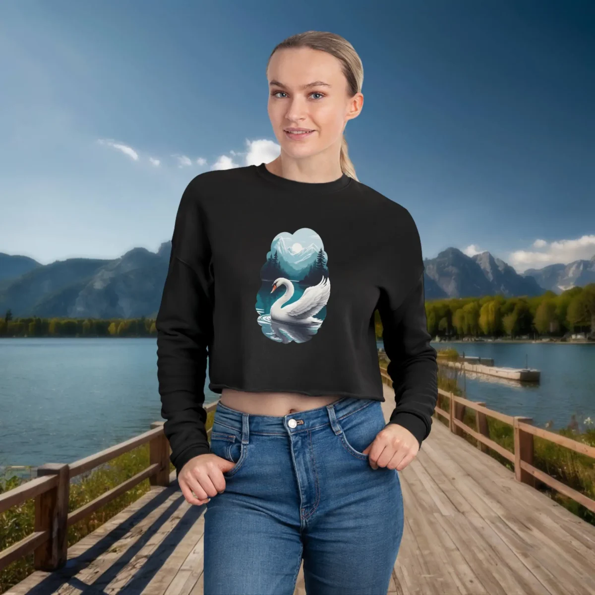 Woman wearing a black cropped sweatshirt with a white swan on a lake design, standing on a wooden dock with a mountain and lake view in the background.