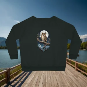 Flat lay of dark green sweatshirt with detailed owl and moon graphic, displayed on a wooden dock by the lake