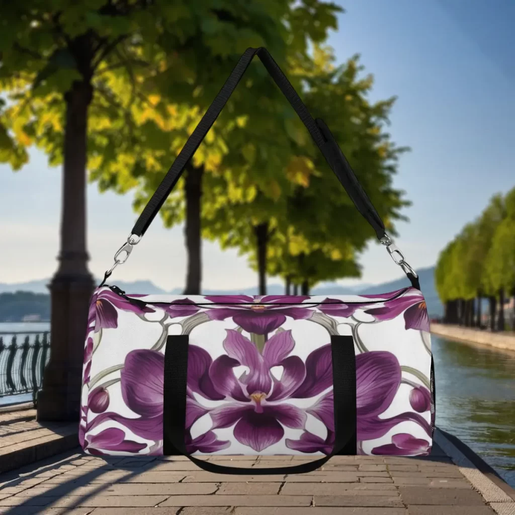 Stylish duffel bag with a purple orchid floral design, displayed outdoors on a scenic walkway with trees and a lake in the background.