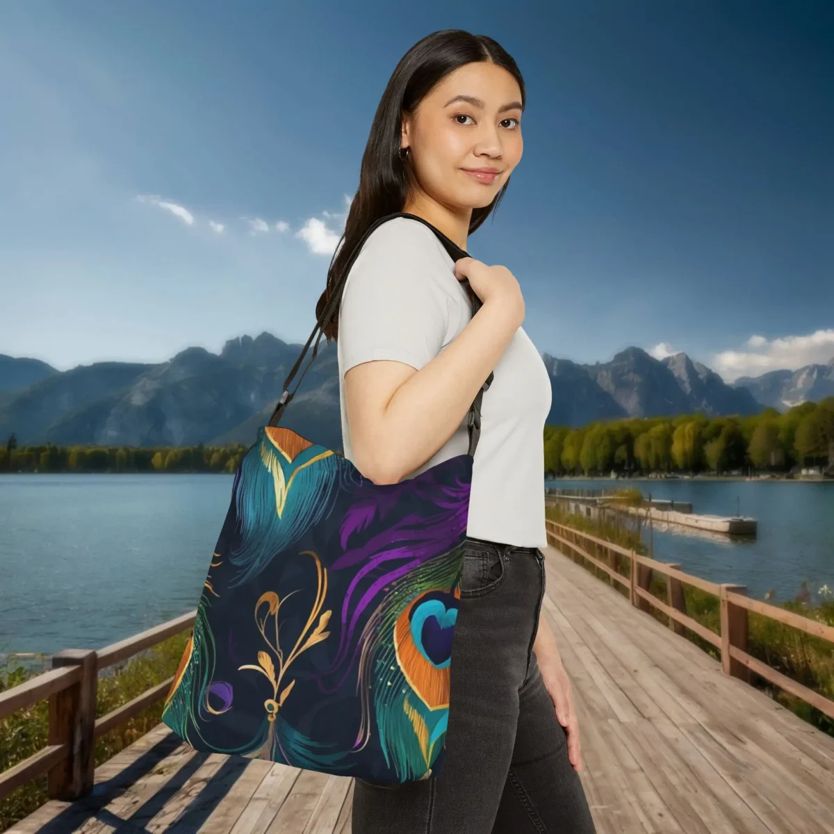 Woman wearing a tote bag with multicolored peacock feather print, standing on a wooden path by a lake and mountains.
