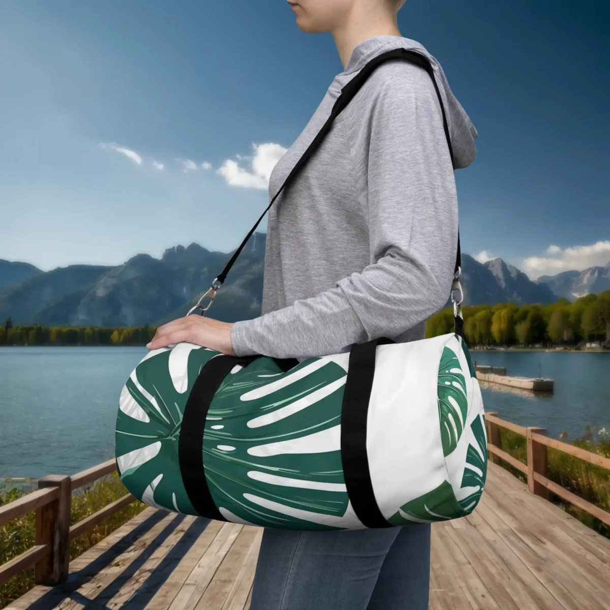 Person carrying a small white duffel bag with green monstera leaf design by a scenic lake.