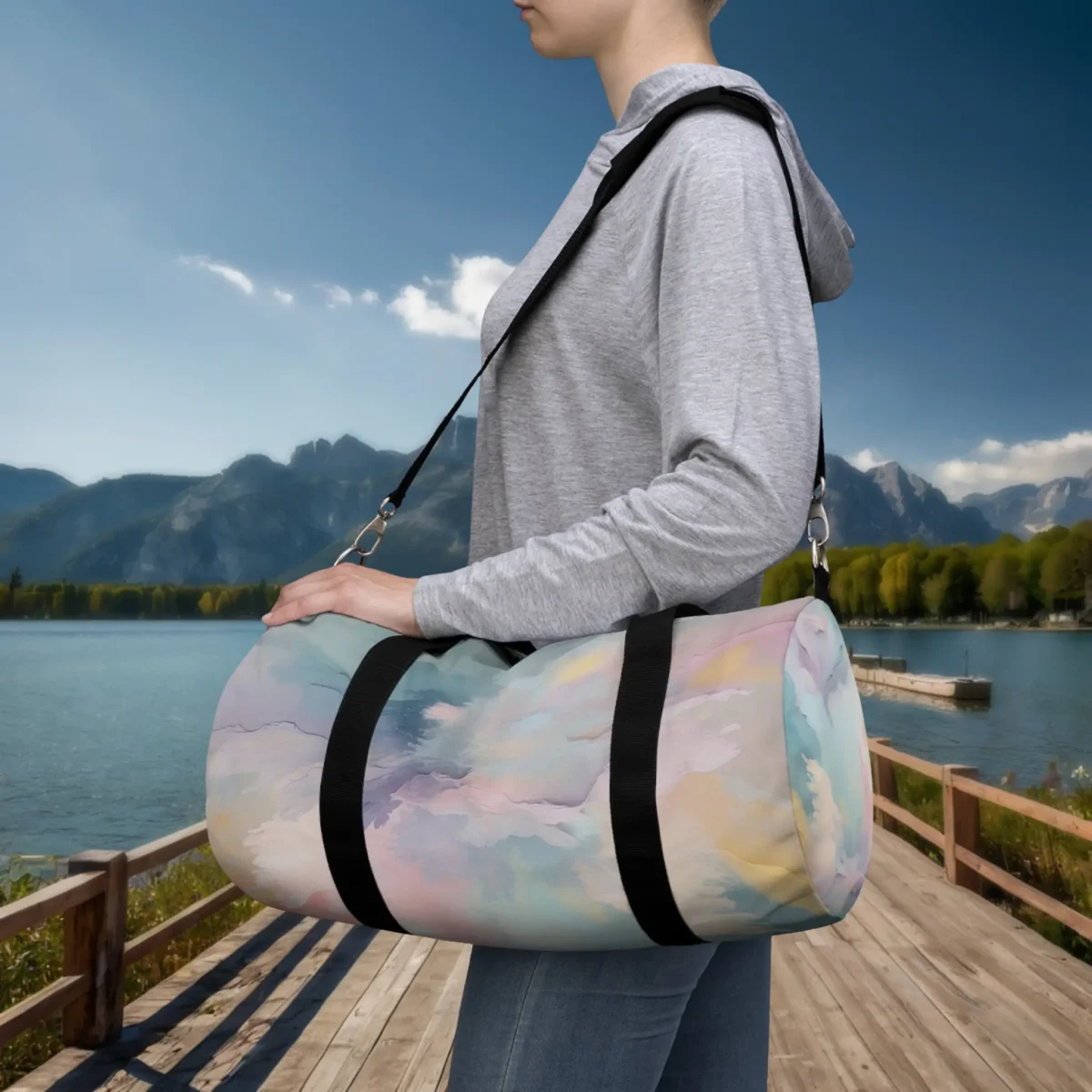 Person carrying a small pastel watercolor duffel bag with black strap, outdoors by a lake and mountain landscape.