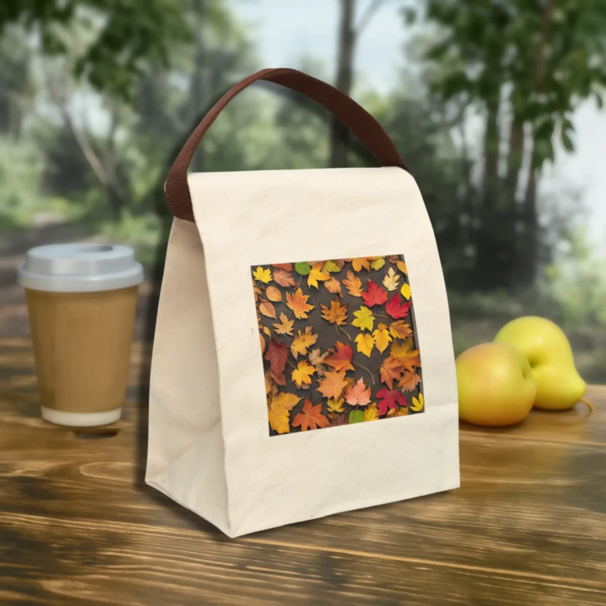 Canvas lunch bag with fall leaves photo print and brown handle, placed on an outdoor wooden table with fruit and coffee cup.