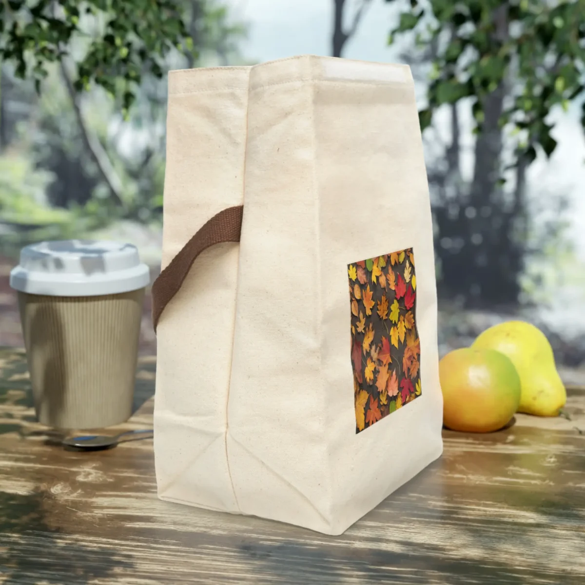 Side view of a cream canvas lunch bag with brown strap and autumn leaves print on the front, placed on a wooden table.