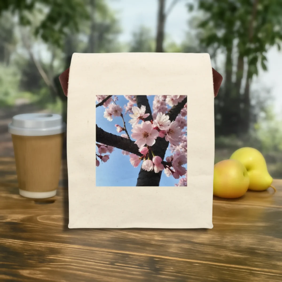 Flat front view of canvas lunch bag featuring a cherry blossom image, displayed on a wooden table with fruit and coffee.