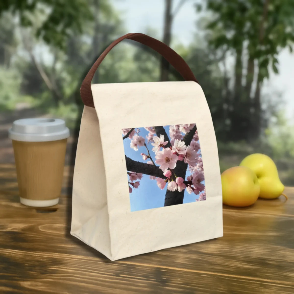 Front angled view of a reusable canvas lunch bag with cherry blossom print and brown handle, displayed on an outdoor table.