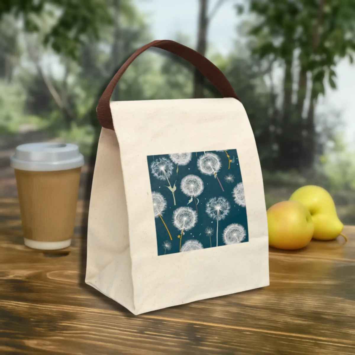 Canvas lunch bag with whimsical white dandelion design and brown strap, styled on an outdoor wooden table with fruit and a coffee cup.