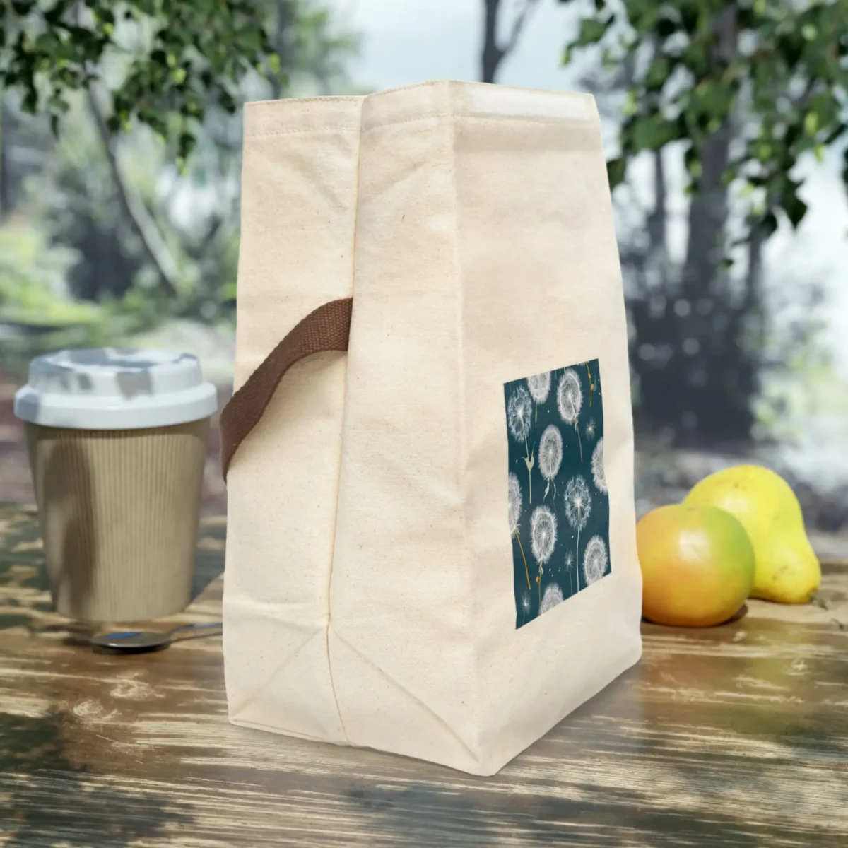 Side view of a cream canvas lunch bag with brown strap and artistic dandelion print on a dark teal background, placed on a wooden outdoor table.
