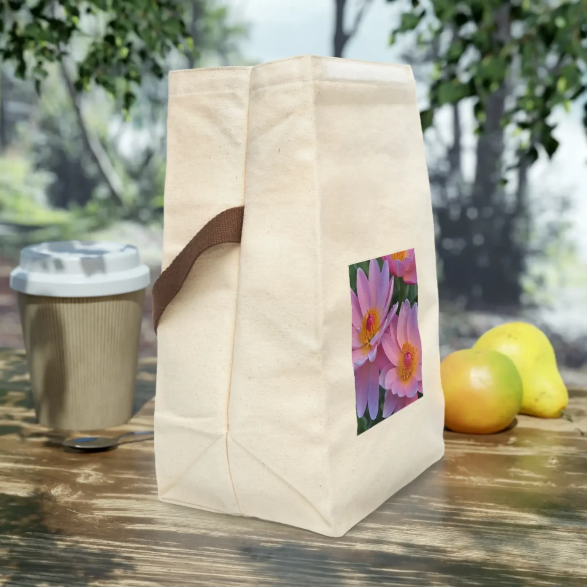 Side view of a cream canvas lunch bag with brown strap and a close-up floral print of pink lotus-like flowers, placed on an outdoor wooden table.