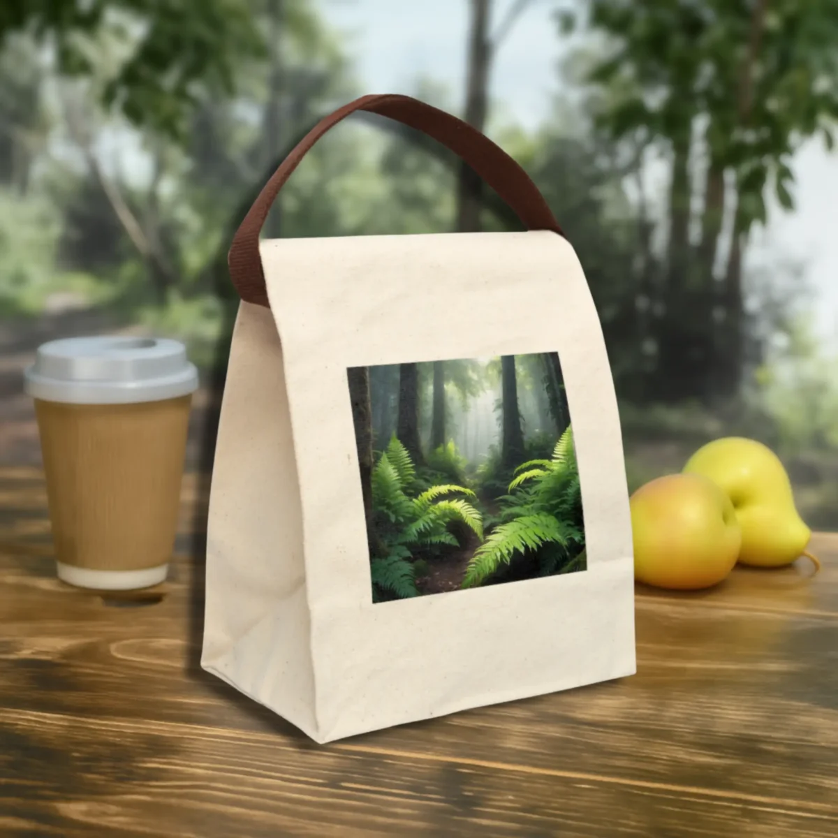 Front angle of a canvas lunch bag with a green forest fern photo and brown handle, styled outdoors with apples and coffee.