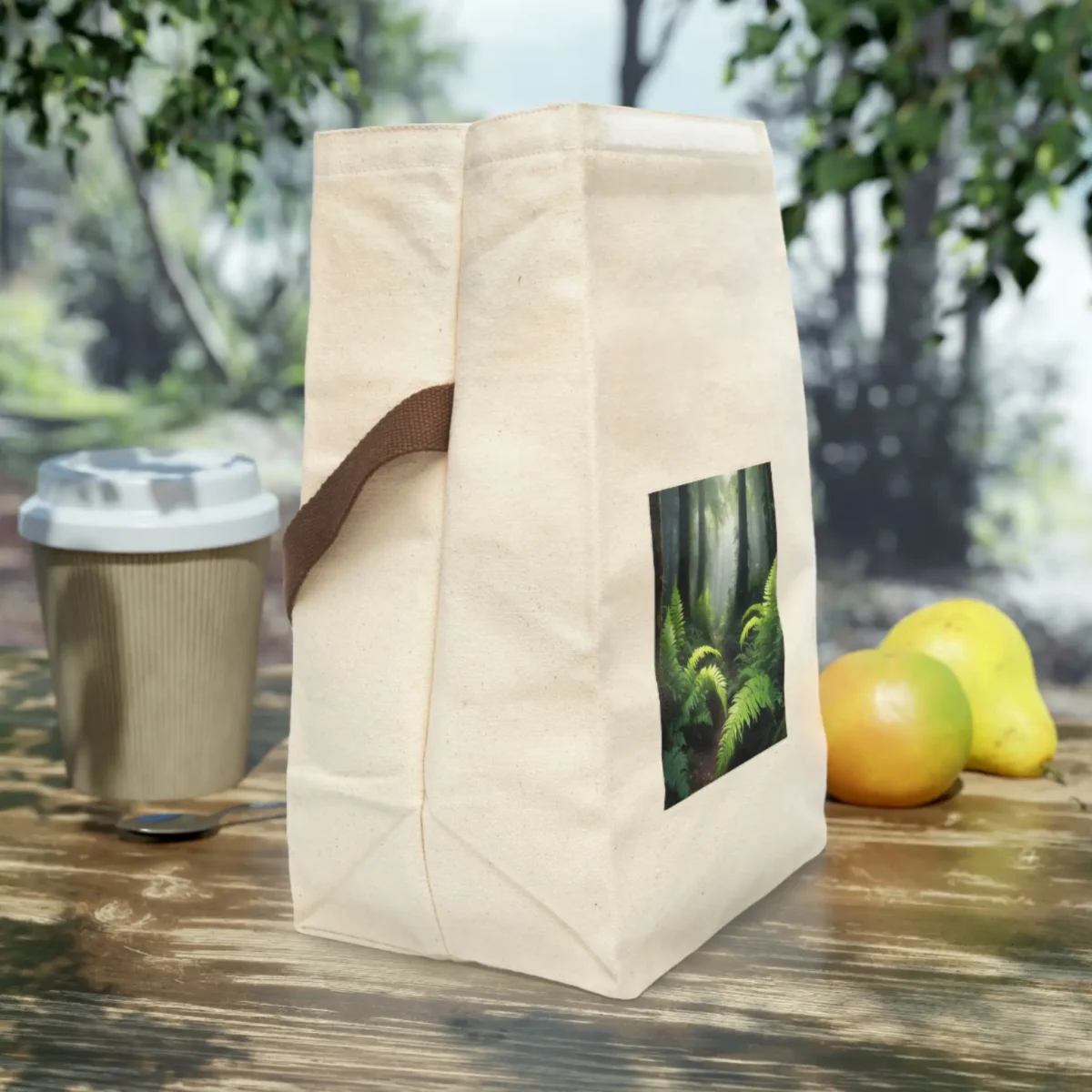 Side view of a cream canvas lunch bag with brown strap and a lush forest fern print, placed on an outdoor wooden table.