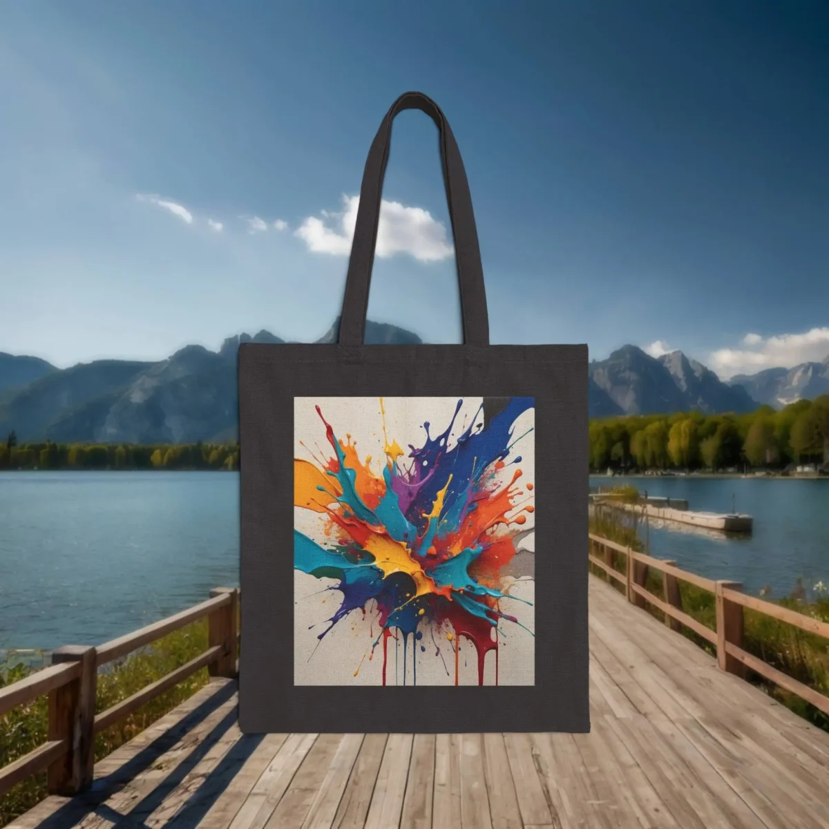 Flat black tote bag with a colorful splatter paint design, placed on a boardwalk by a calm lake with mountain scenery.
