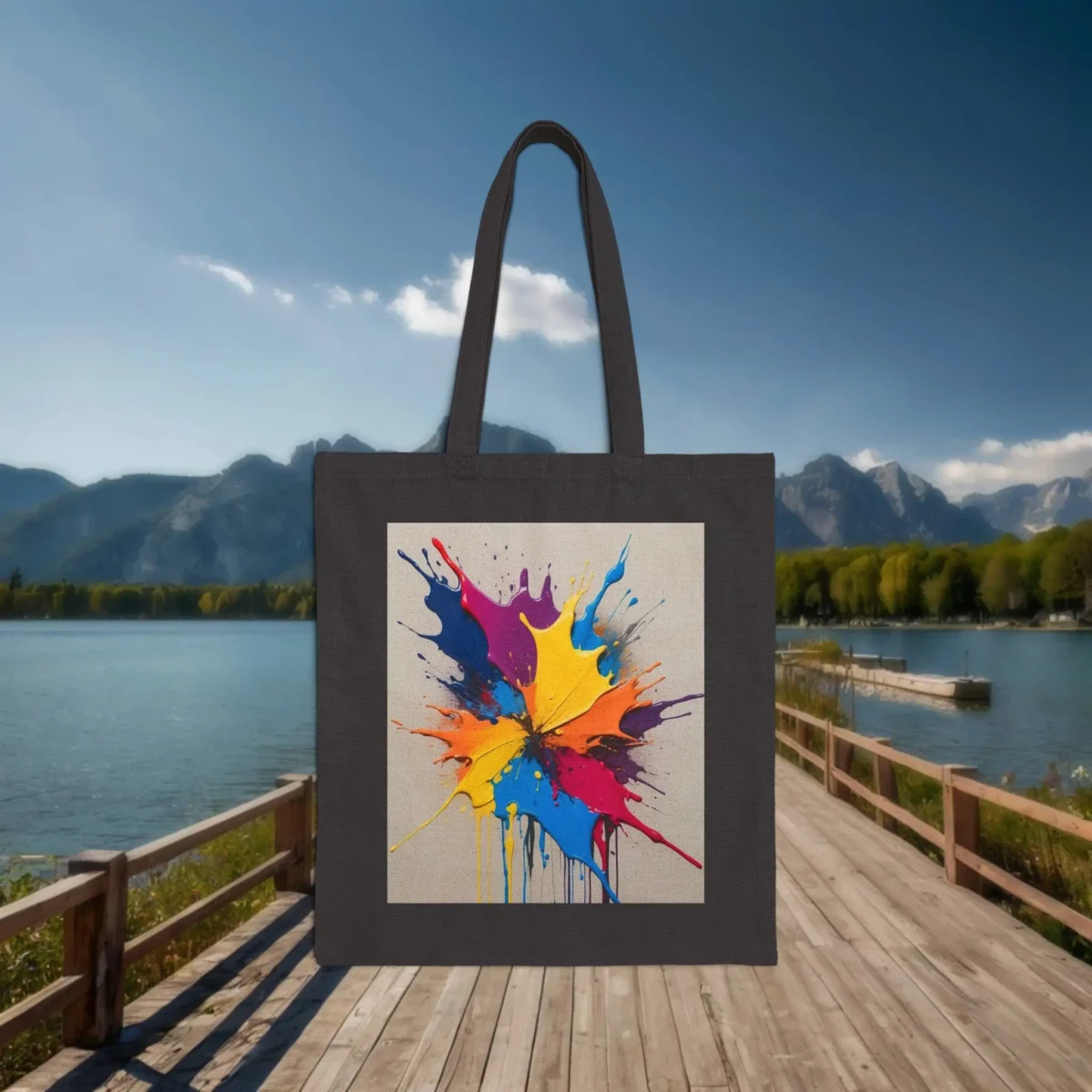 Flat black tote bag with a colorful splash of dripping paint in a radial pattern, displayed on a dock near a lake.