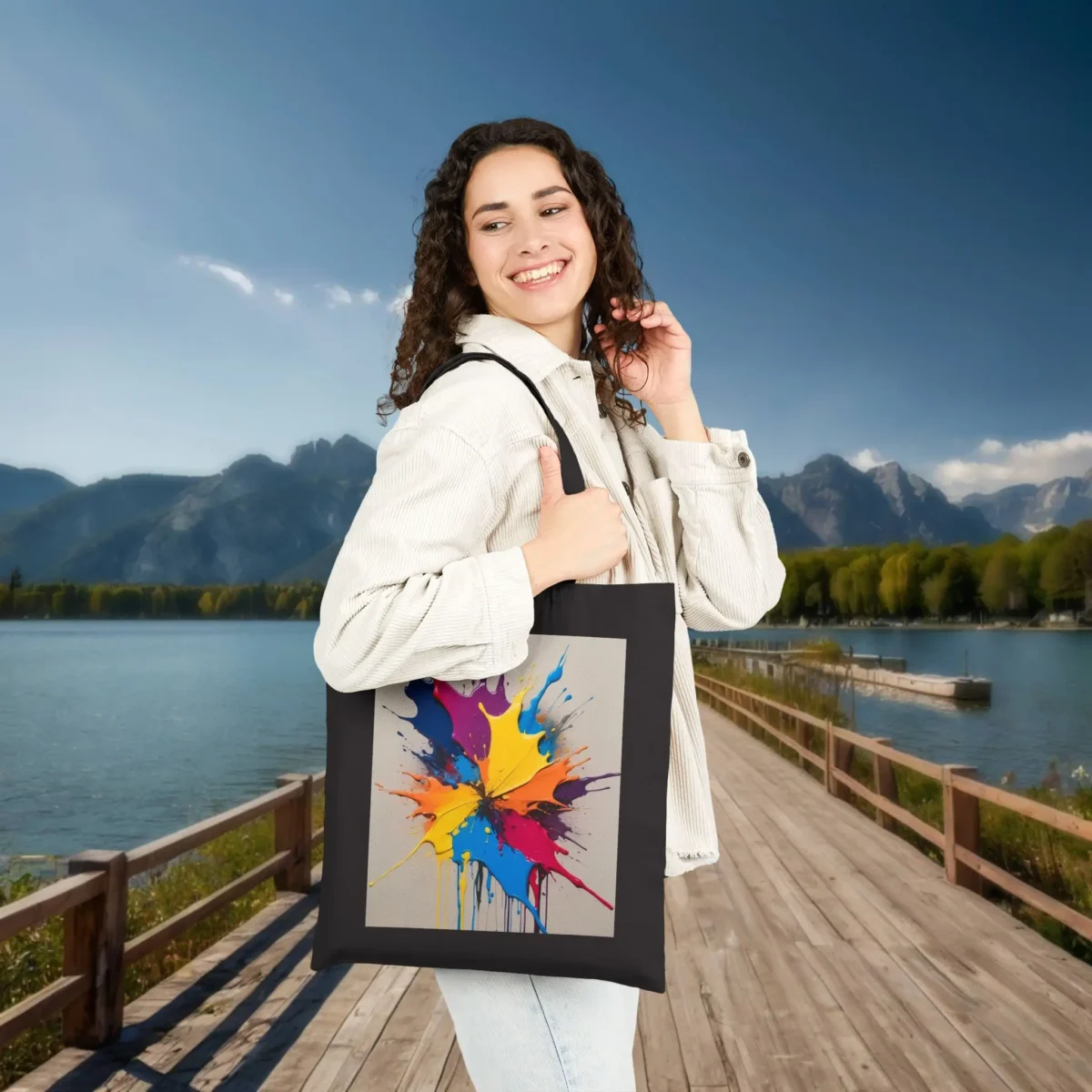 Woman carrying a black tote bag with a vivid splash of colorful paint in yellow, red, blue, and pink tones, standing on a scenic boardwalk.