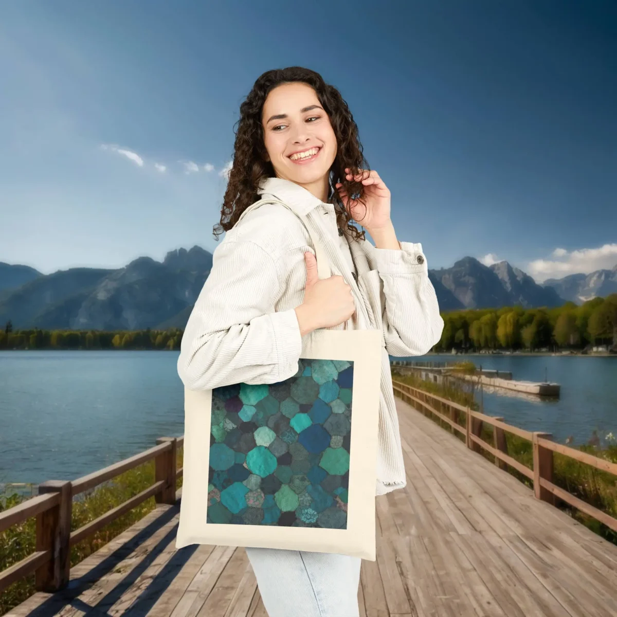Woman smiling while carrying a natural-colored tote bag with a green and teal hexagon mosaic print in a mountain lakeside setting.