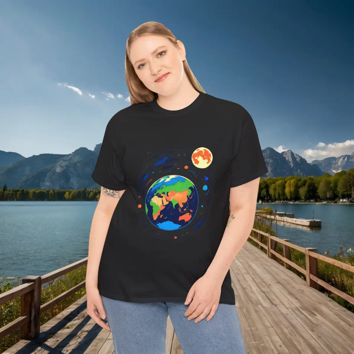 Space-themed black t-shirt with Earth and Moon – women’s fit Woman in a black t-shirt with a colorful Earth and Moon space design, posing on a dock with a scenic mountain lake view.