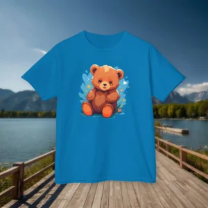 Front view of a light blue kids’ t-shirt featuring a cartoon baby bear with a soft expression, surrounded by heart and star decorations, displayed on a dock by a lake.