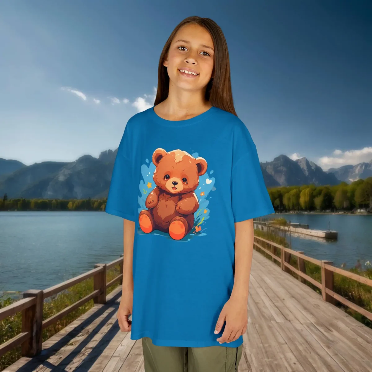 Smiling girl wearing a light blue t-shirt with a cute cartoon baby bear surrounded by blue and yellow hearts, standing by a peaceful lake.