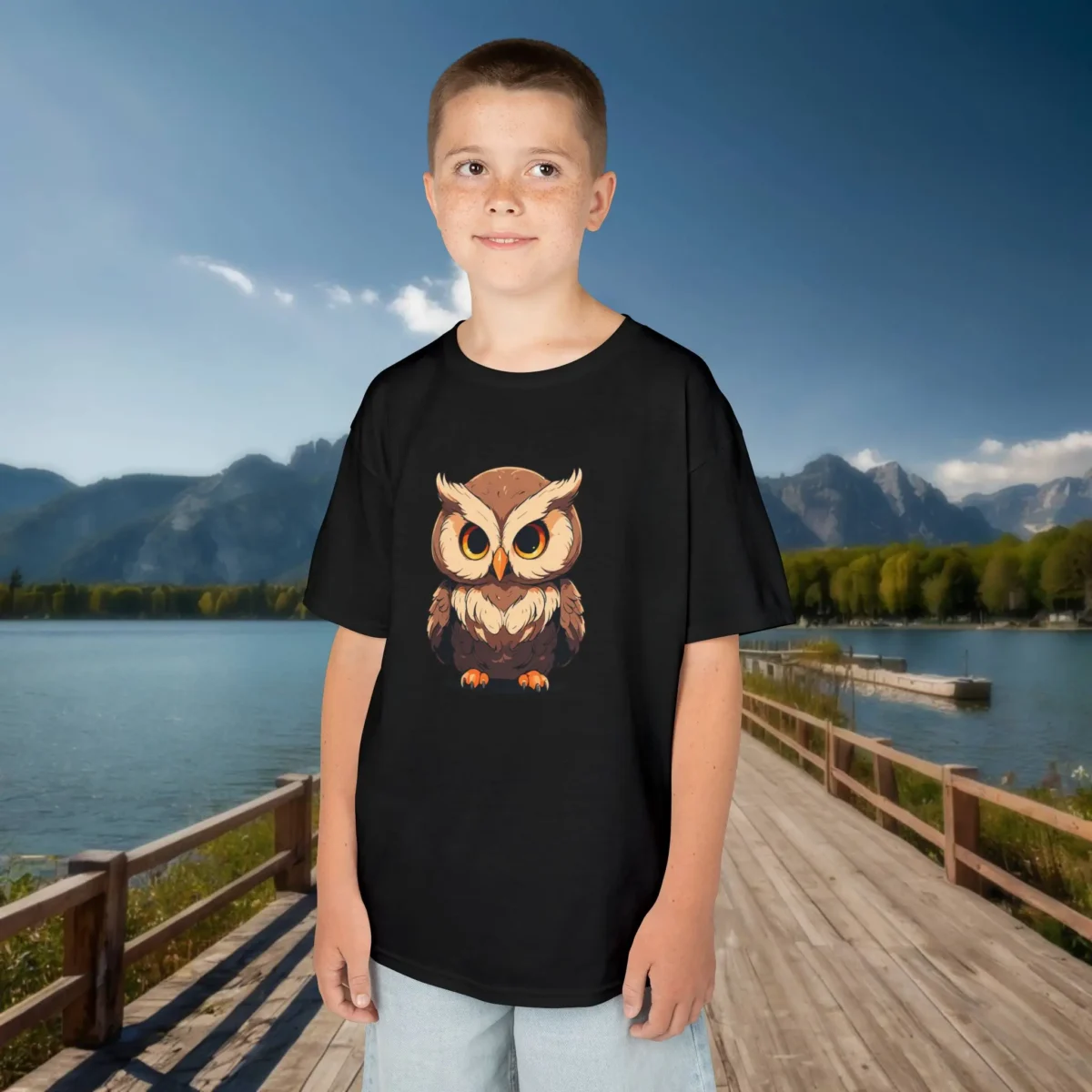Smiling boy wearing a black t-shirt with a cartoon baby owl design featuring intense orange eyes, standing near a scenic lake.