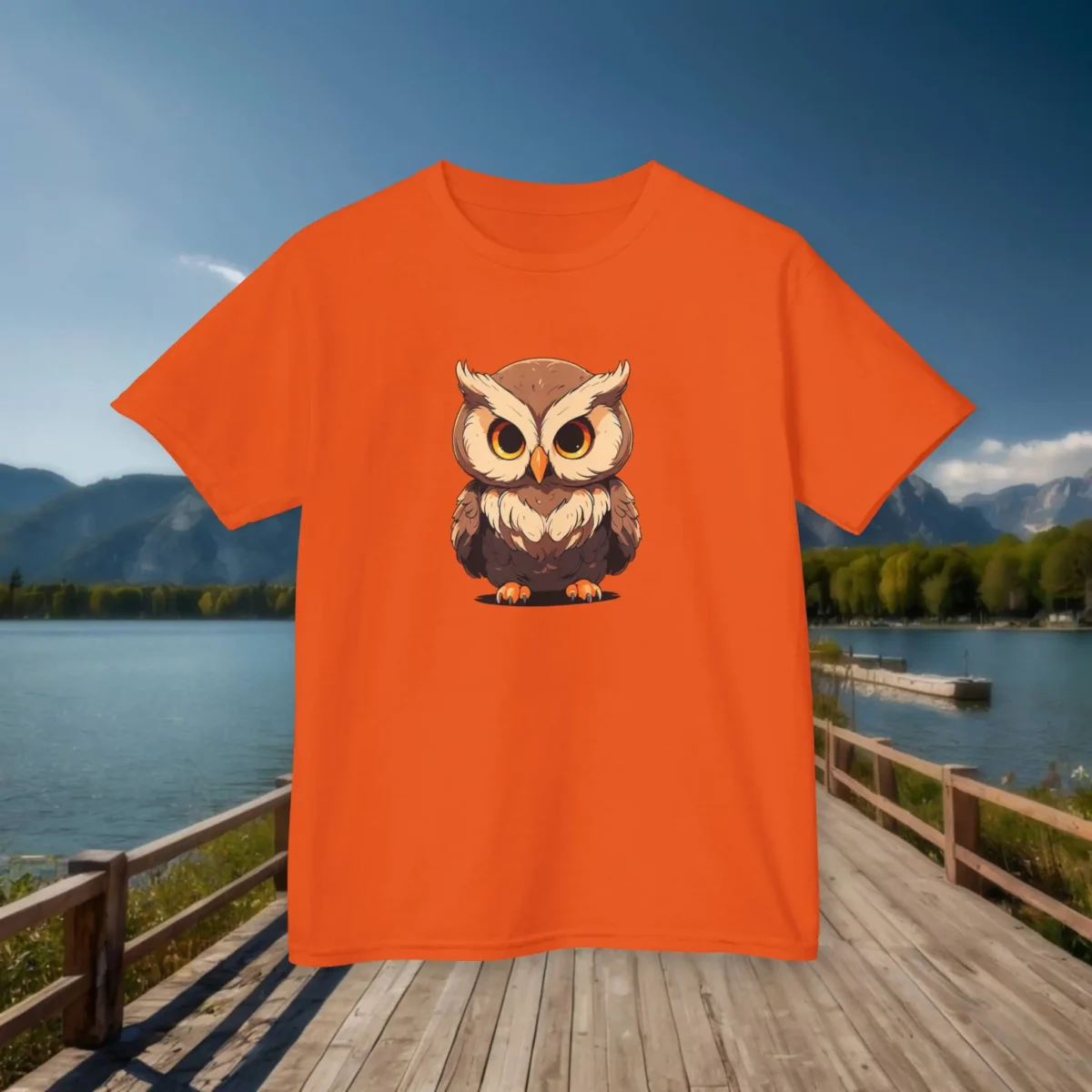 Front view of an orange kids' t-shirt with a cartoon baby owl featuring large eyes and fluffy feathers, displayed on a boardwalk by a lake.