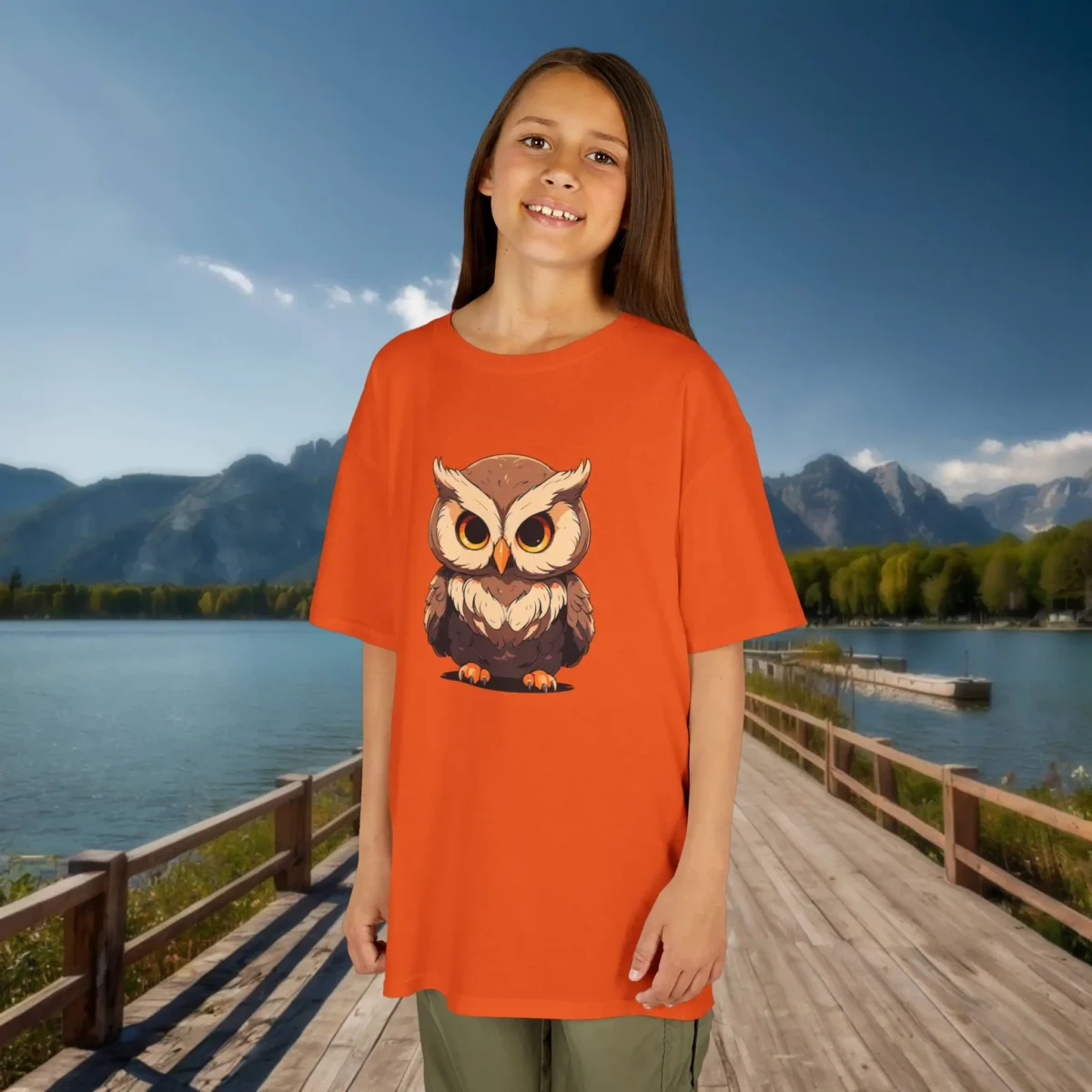 Girl wearing an orange t-shirt with a cute cartoon baby owl design, standing on a wooden dock by the lake.