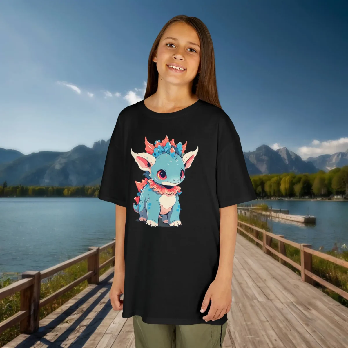 Girl modeling a black T-shirt with a cute baby Styracosaurus illustration, standing on a dock by a mountain lake.