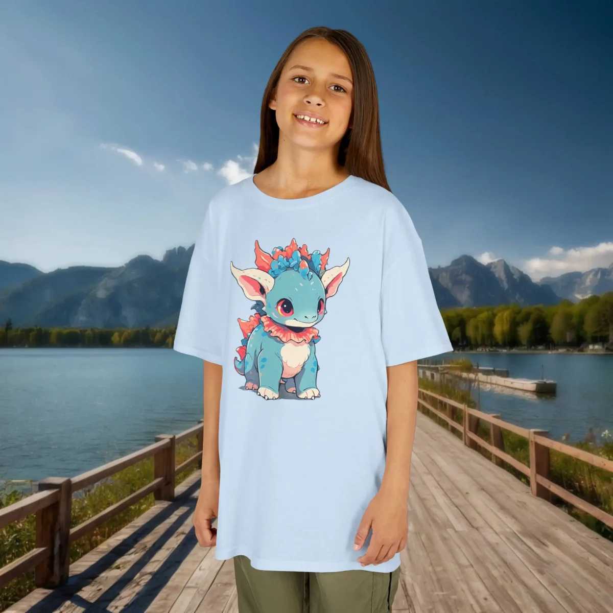 Girl wearing a light blue T-shirt featuring a cute baby Styracosaurus cartoon, standing on a wooden bridge with a lake and mountains in the background.