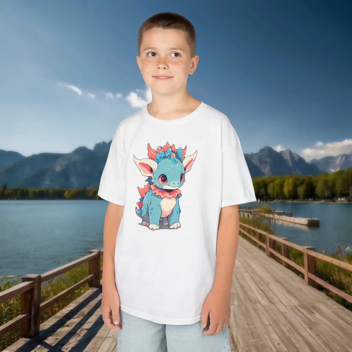 Boy wearing a white T-shirt with a baby Styracosaurus cartoon design, standing outdoors on a wooden dock with mountains in the background.
